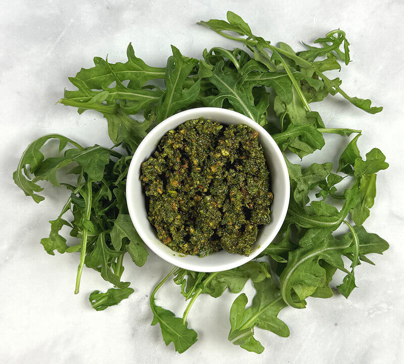 An overhead shot of arugula pistachio pesto in a white bowl on a bed of arugula.