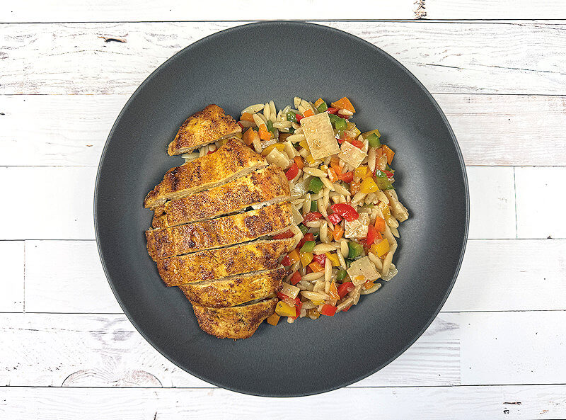 Dark grey plate with bell pepper orzo salad and a cooked chicken breast on top.