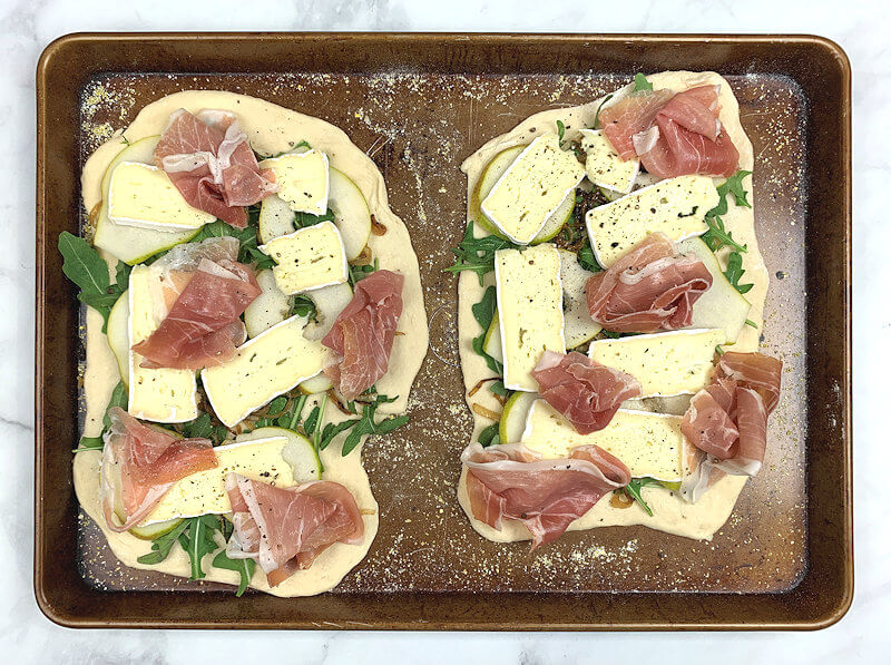 Two small pizzas ready to go into the oven. Toppings are caramelized onions, Prosciutto, Brie cheese and slices of pear.