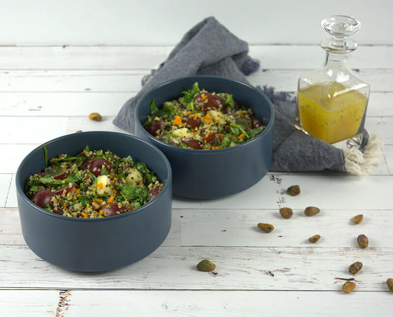 Two dark blue bowls filled with quinoa salad with grapes. Orange dressing is in background in glass jar.