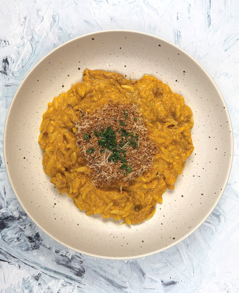 Cream colored bowl with Creamy Carrot & Chicken Orzo. Garnished with toasted breadcrumbs and parsley