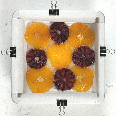Step 5 - In a glass jar or bowl, combine water and sugar and heat in the microwave, until sugar is completely dissolved. Allow to cool for a few minutes. In the meantime, peel oranges and slice super thin, making use of a sharp mandoline or cut by hand. Pour the syrup into a 6x6 baking dish, lined with parchment paper. Arrange the orange slices at the bottom of the baking dish, in the syrup.