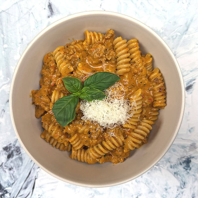 Bowl with roasted vegetable pasta with sausage. Topped with Parmigiano Reggiano cheese and basil.