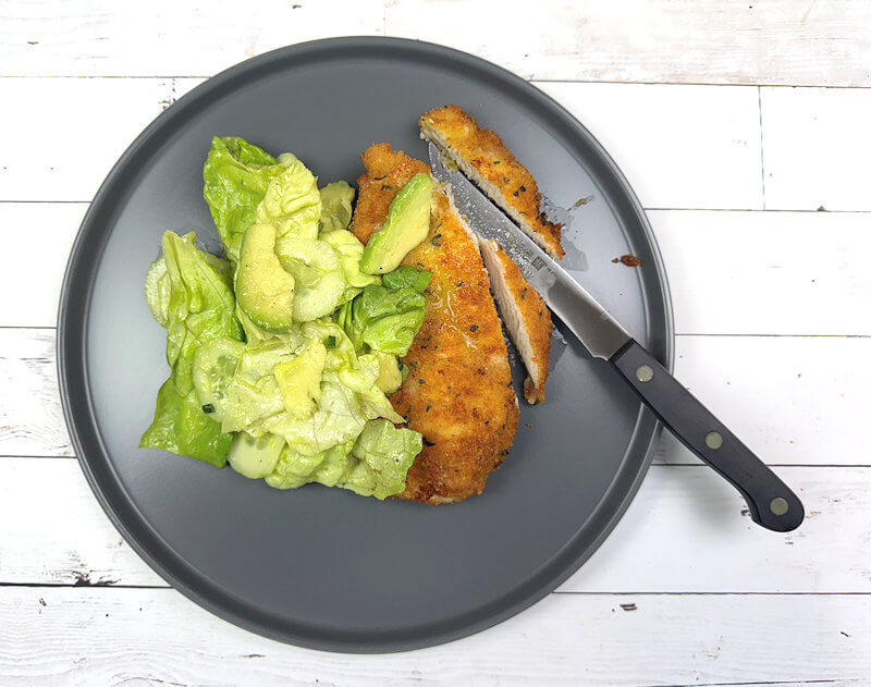 Dark grey plate with chicken Milanese with Butter Lettuce salad. Knife with a few cut in the chicken.