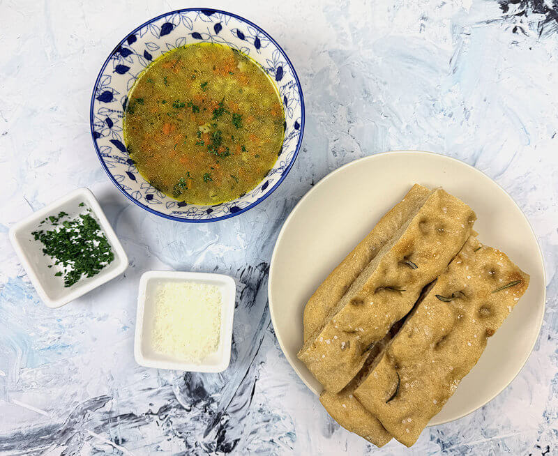 Soup served in a bowl with small plates with focaccia, parsley and grated Parmigiano cheese