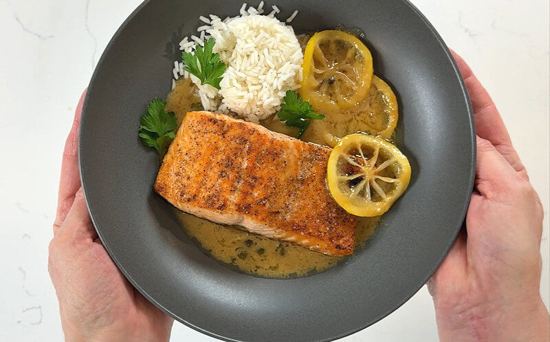 Two hands serving a plate of salmon piccata with miso, rice, lemon slices and some parsley leaves.