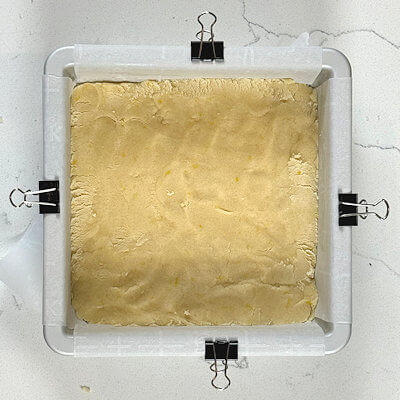 Step 4 - With your finger, press 2/3 of the dough into a 6x6 baking dish, lined with parchment paper. I always cut it 2 inches longer and use binder clips to keep the parchment paper in place.