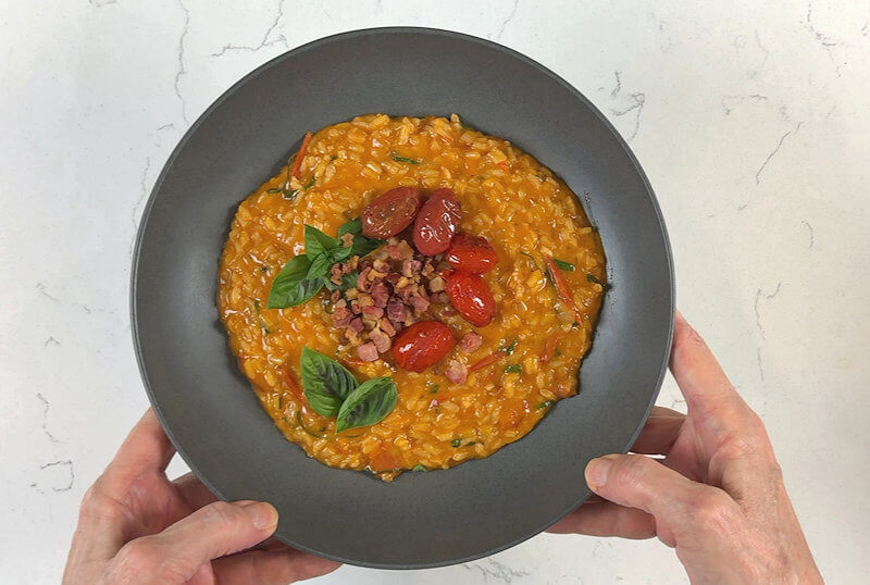 Grey plate with tomato risotto, garnished with the crispy pancetta, tomatoes and basil leaves.